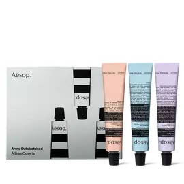 Tre tubi di balsamo per le mani Aesop color pesca, azzurro pallido e lilla poggiano sui rispettivi tappi neri accanto a una scatola illustrata grigio argento.