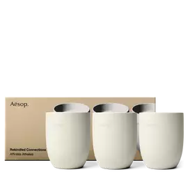 Tre recipienti color crema per candele Aesop sono allineati davanti a una scatola dorata stampata con immagini delle candele corrispondenti.