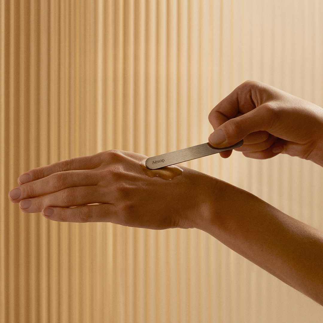 Una persona usa una spatola Aesop per applicare un prodotto per la cura della pelle sul dorso della mano.
