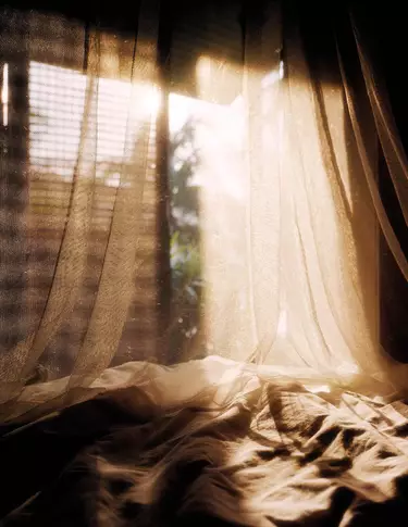 Tende leggere drappeggiate su tessuto stropicciato accanto a una finestra illuminata dal sole, in calde tonalita beige, con ombre morbide e pieghe traslucide.