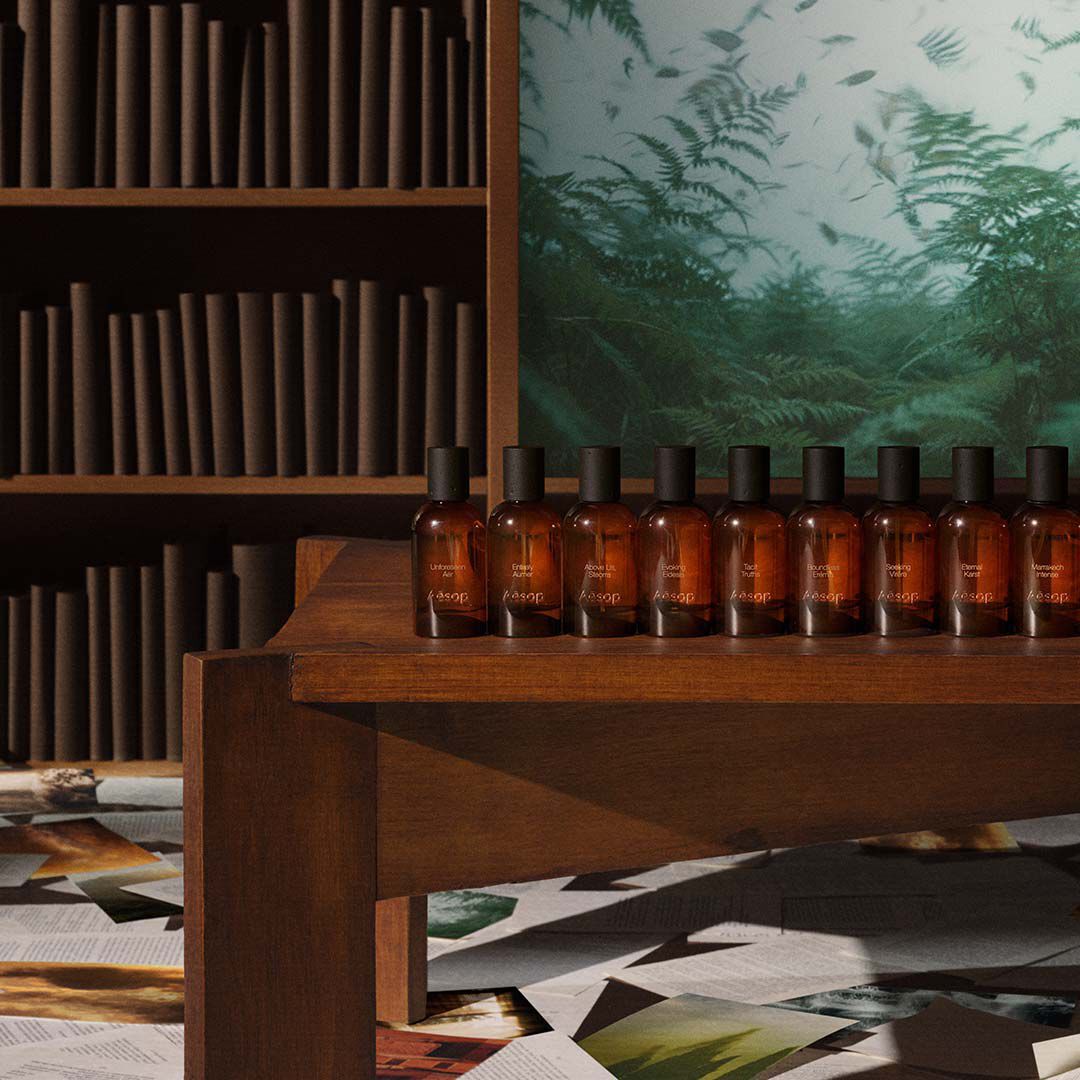 Una fila di flaconi di profumo in vetro ambrato Aesop esposti su un ripiano di legno davanti a una libreria e a un'opera d'arte verde.