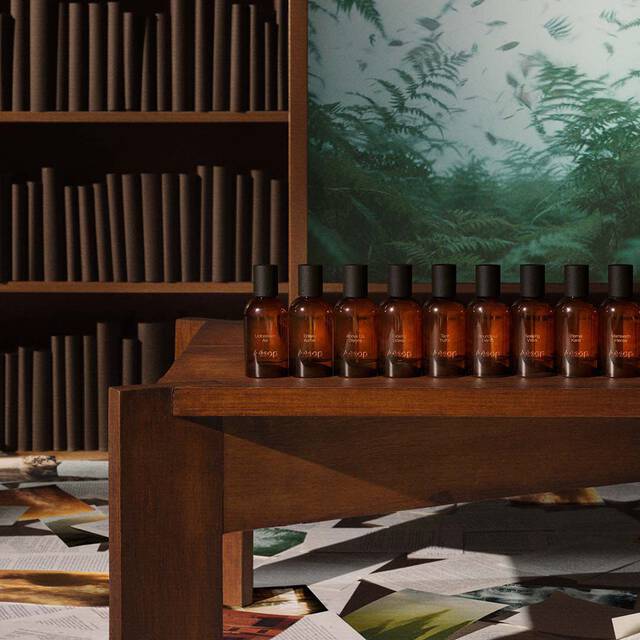 Una fila di flaconi di profumo in vetro ambrato Aesop esposti su un ripiano di legno davanti a una libreria e a un'opera d'arte verde.