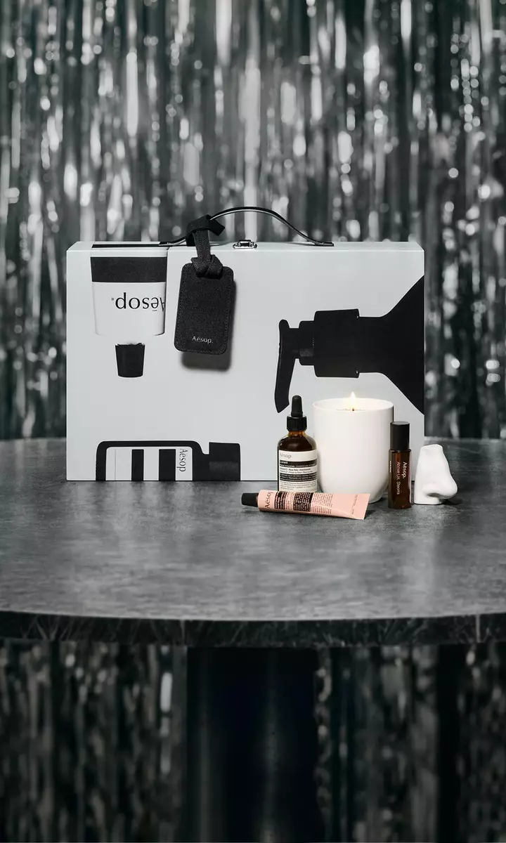 Confezione regalo e prodotti skincare Aesop su un tavolo, con uno sfondo festivo dalle luci soffuse.