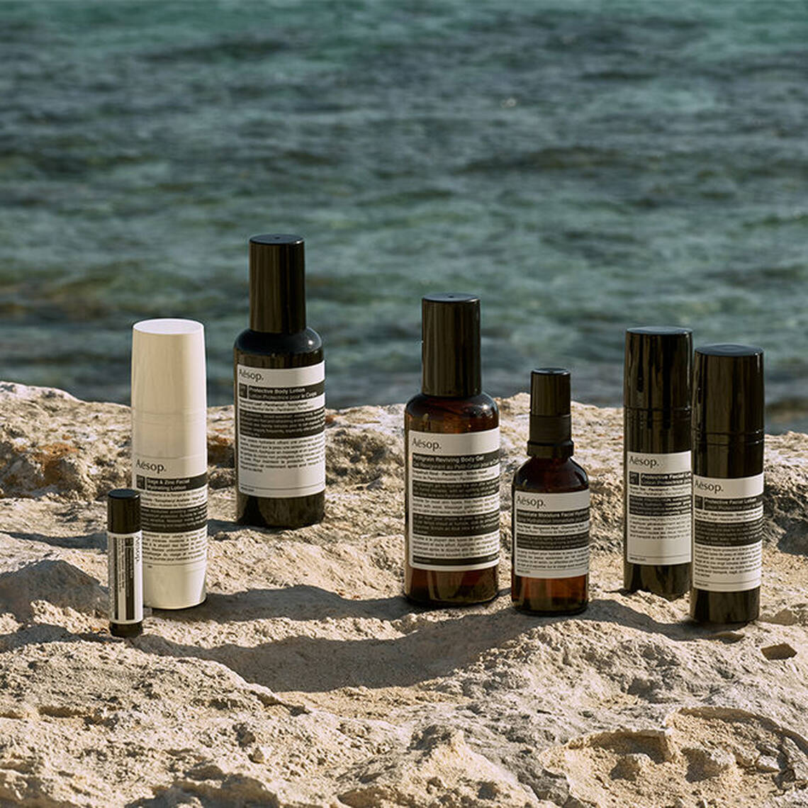 Une selection soignee d'allies protecteurs solaires Aesop presentes sur une roche cotiere au bord de la mer.