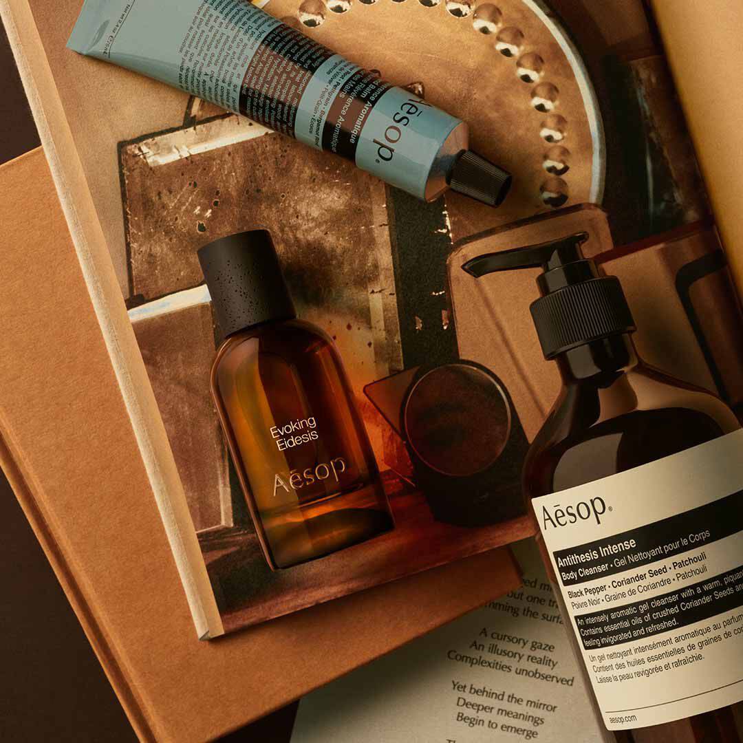 Flacone Aesop Evoking Eidesis con flacone a pompa e tubo blu, disposti su fogli e immagini di un vassoio a specchio.