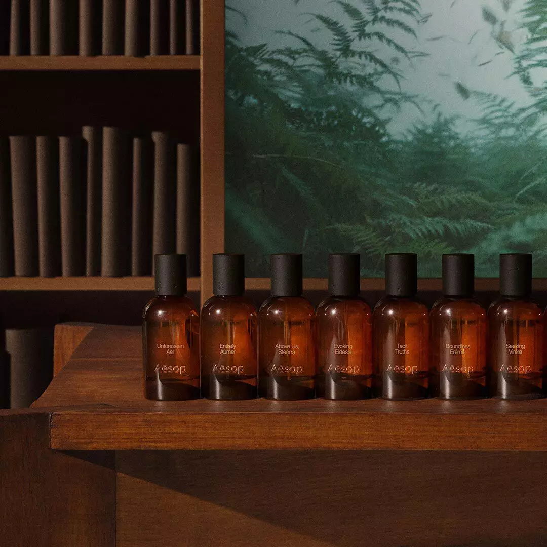 Fila di flaconi ambrati di fragranze Aesop su un tavolo di legno, accanto a scaffali e all’immagine di una felce verde.