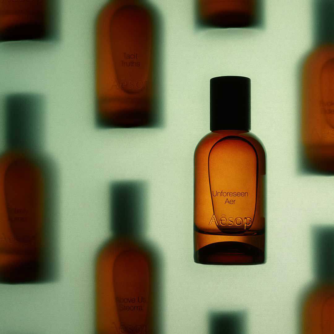 Flacone ambrato di fragranza Aesop Unforeseen Aer a fuoco, davanti a flaconi ambrati sfocati su uno sfondo verde pallido.