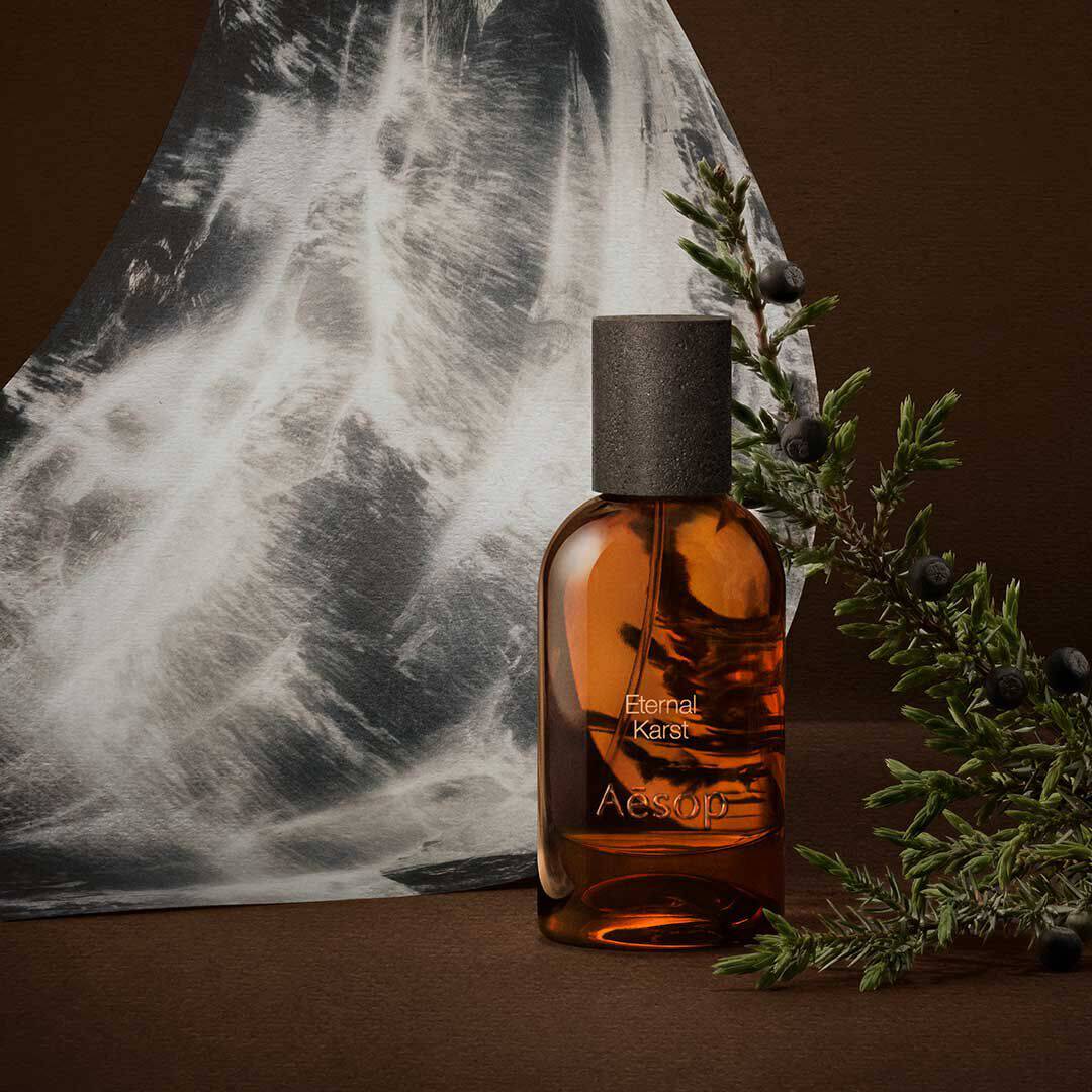 Flacone ambrato di fragranza Aesop accanto a rametti di rosmarino e a un’immagine monocromatica d’acqua, su una superficie marrone scuro.