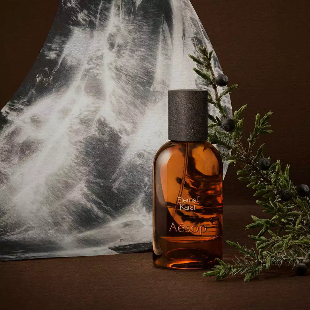 Flacone ambrato di fragranza Aesop accanto a rametti di rosmarino e a un’immagine monocromatica d’acqua, su una superficie marrone scuro.