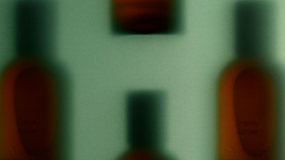 Flaconi di fragranze ambrate dai contorni sfumati, con tappi scuri su uno sfondo verde pallido, a creare un motivo orizzontale ripetuto e indistinto.