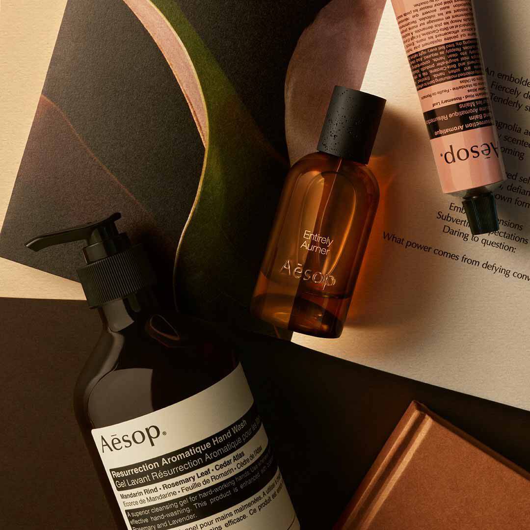 Flacone Aesop Entirely Aurner con flacone a pompa e tubo color pesca, disposti su fogli e forme scure curve.