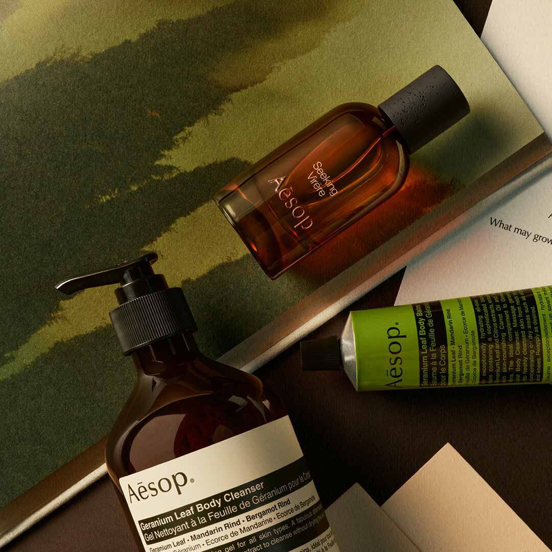 Flacone Aesop Seeking Virere con flacone a pompa e tubo verde, disposti su fogli con immagini dipinte nei toni del verde.
