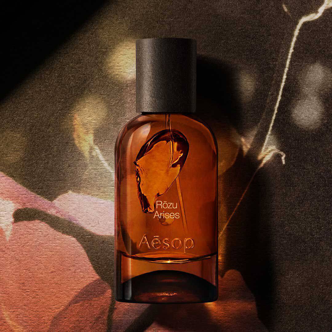 Flacone ambrato di fragranza Aesop Rozu Arises su un’immagine floreale scura, con luce calda e ombre.