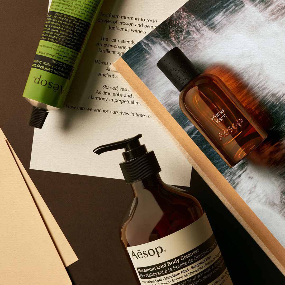 Flacone Aesop Eternal Karst con flacone a pompa e tubo verde, disposti su fogli con immagini di acqua schiumosa.