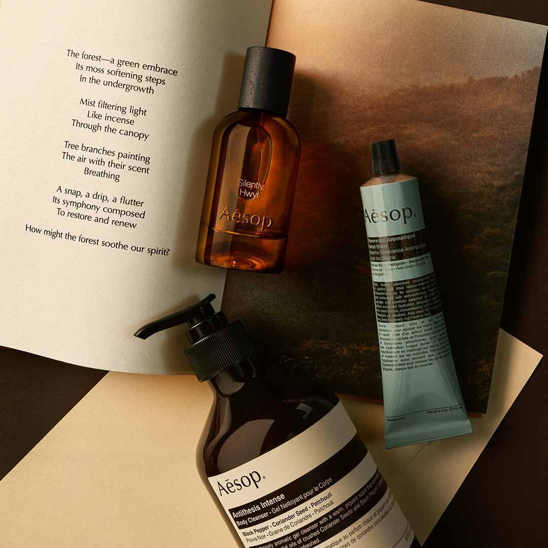 Flacone Aesop Silently Hwyl con flacone a pompa e tubo blu, disposti su fogli con un’immagine di paesaggio.