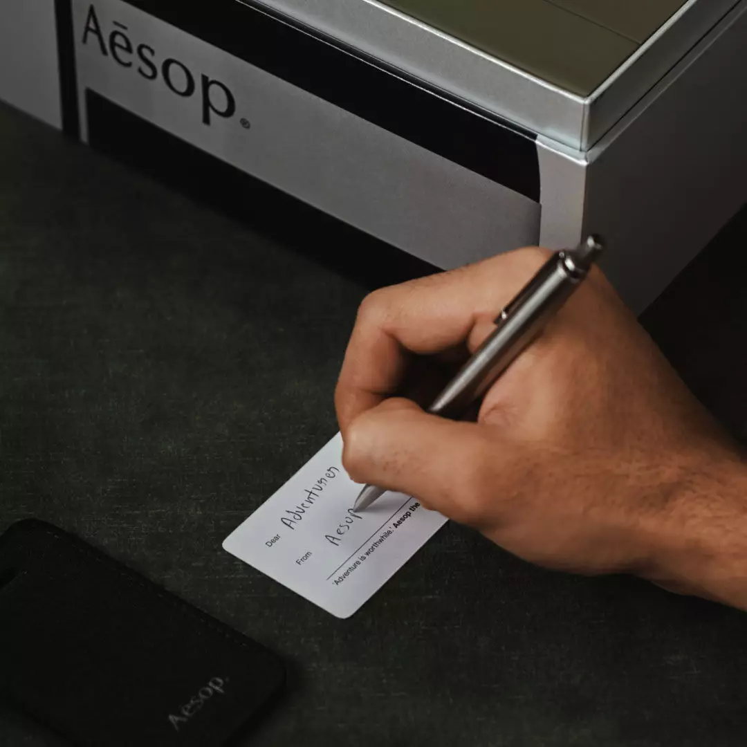 Una mano scrive su un piccolo biglietto bianco accanto a un’etichetta per bagagli Aesop nera e a un astuccio Aesop argentato, su tessuto verde scuro.