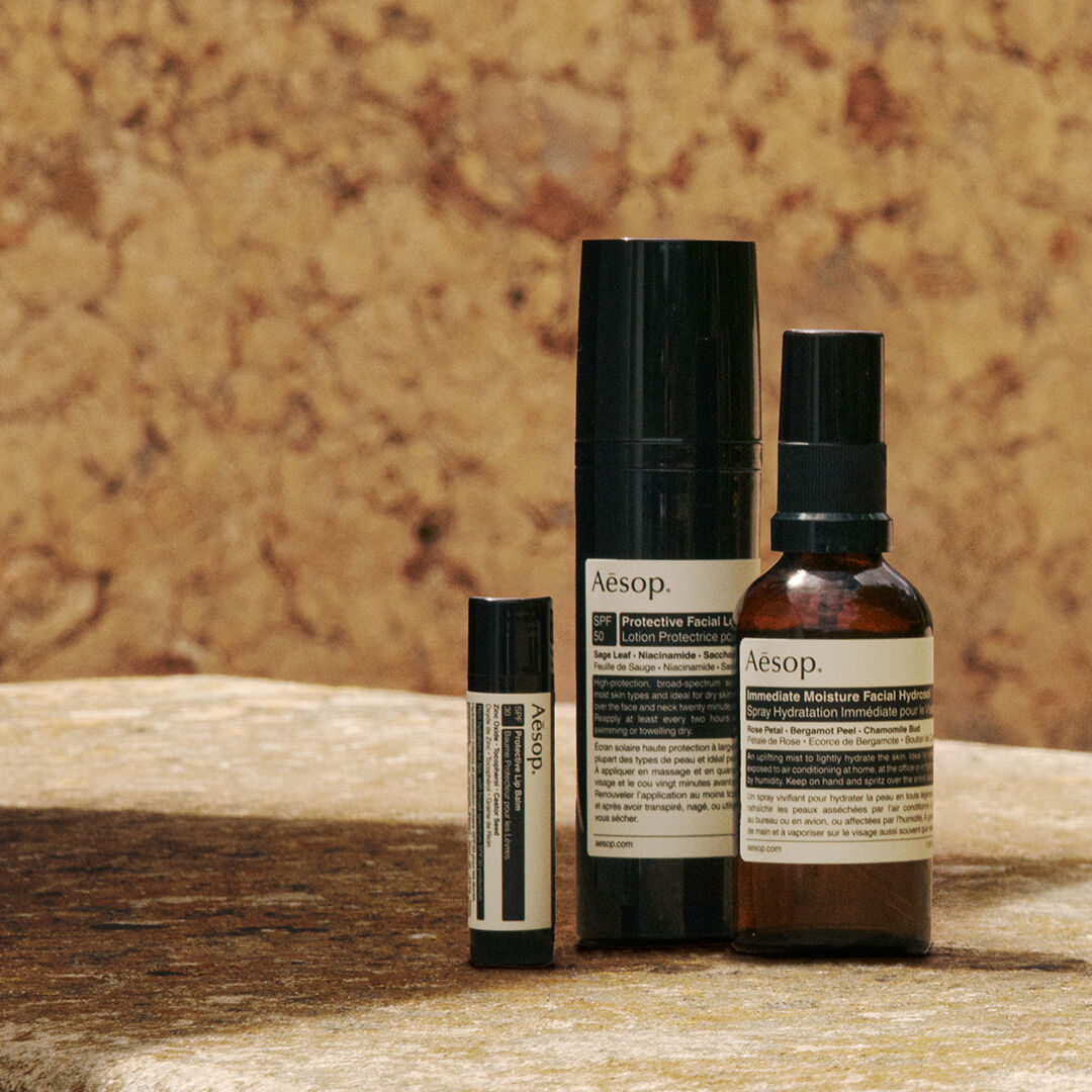 Un'accurata selezione di formulazioni per la protezione solare Aesop disposte su una superficie in pietra illuminata dal sole, contro una parete dalle tonalita calde.
