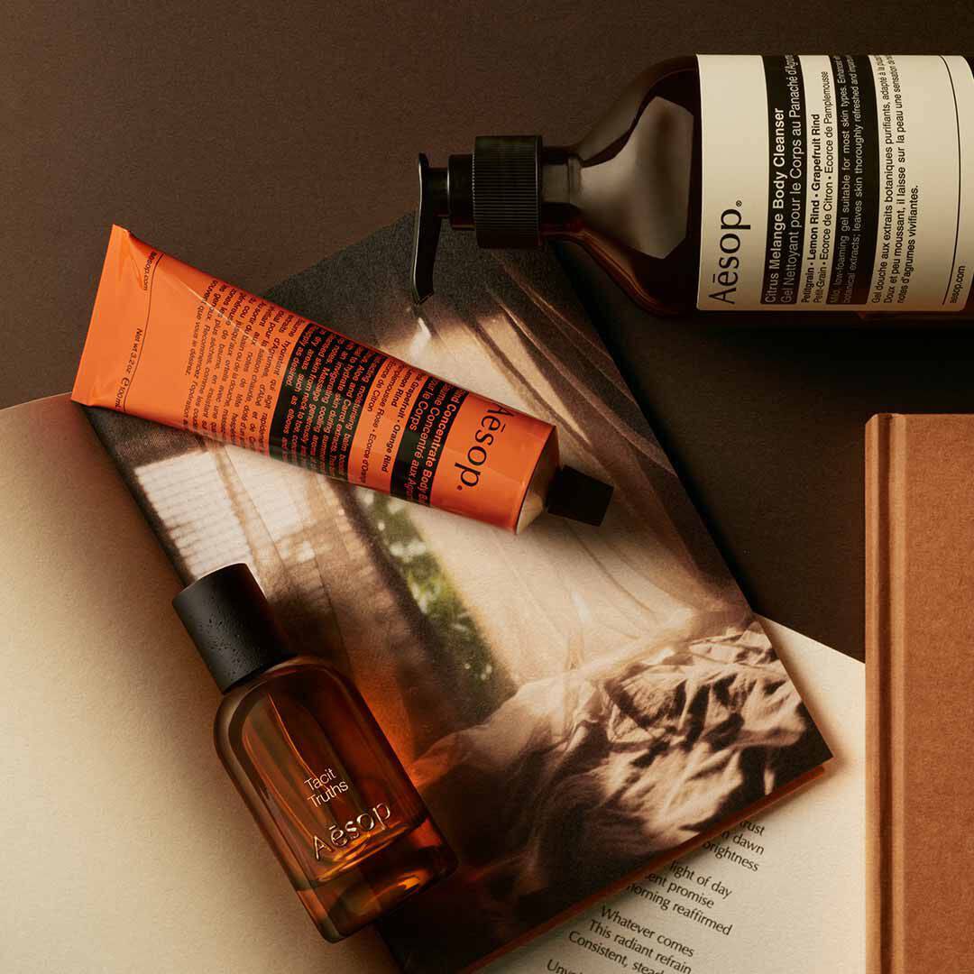 Flacone Aesop Tacit Truths con flacone a pompa e tubo arancione, disposti su fogli con un’immagine monocromatica.
