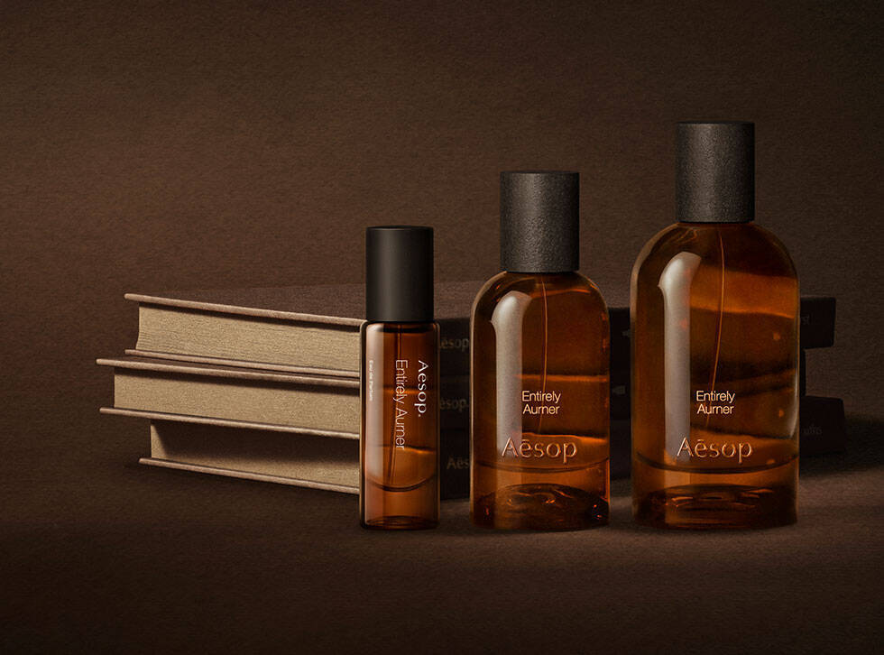 Tre flaconi ambrati di fragranze Aesop disposti accanto a libri color crema impilati su una superficie marrone scuro.