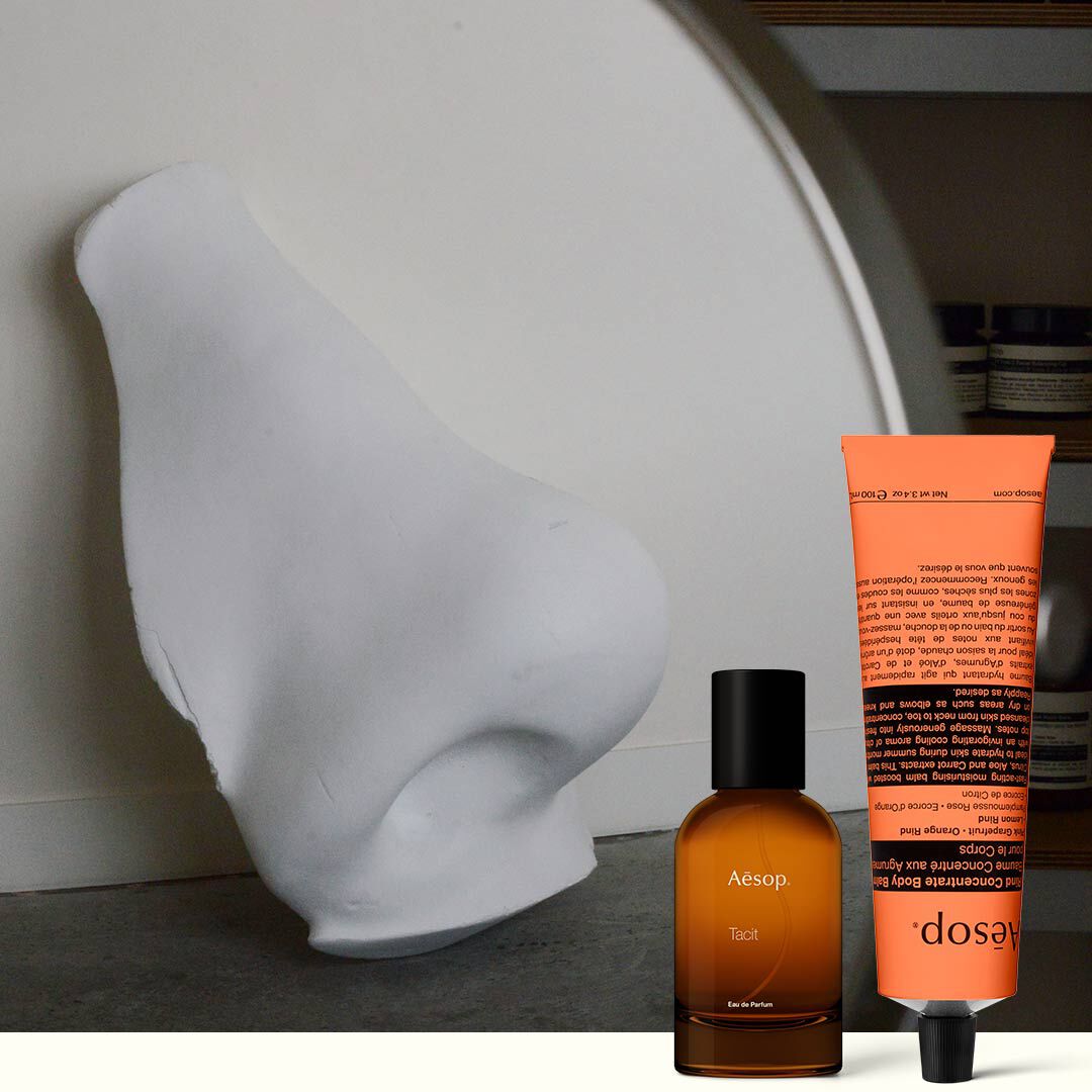 Flacone di profumo ambrato e crema mani color corallo accanto a una scultura bianca