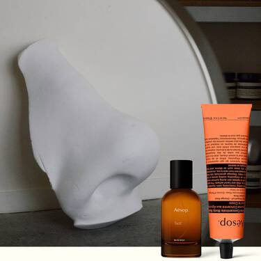 Flacone di profumo ambrato e crema mani color corallo accanto a una scultura bianca