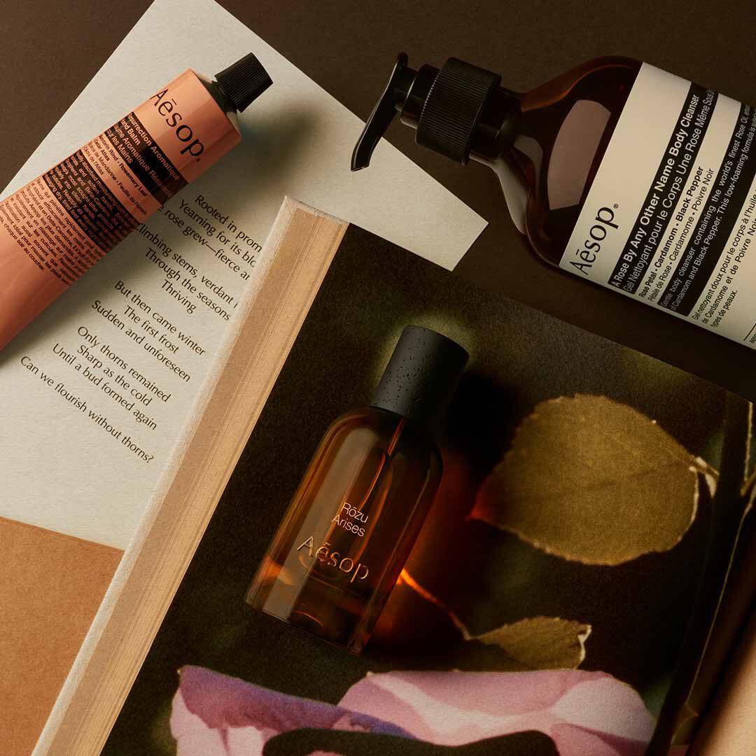 Flacone Aesop Rozu Arises con flacone a pompa e tubo color pesca, disposti su fogli con petali rosa e una foglia.