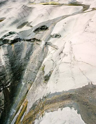 Superficie minerale in pietra chiara, con venature bianche, grigie e antracite, texture stratificate e una frattura diagonale che attraversa l’inquadratura.