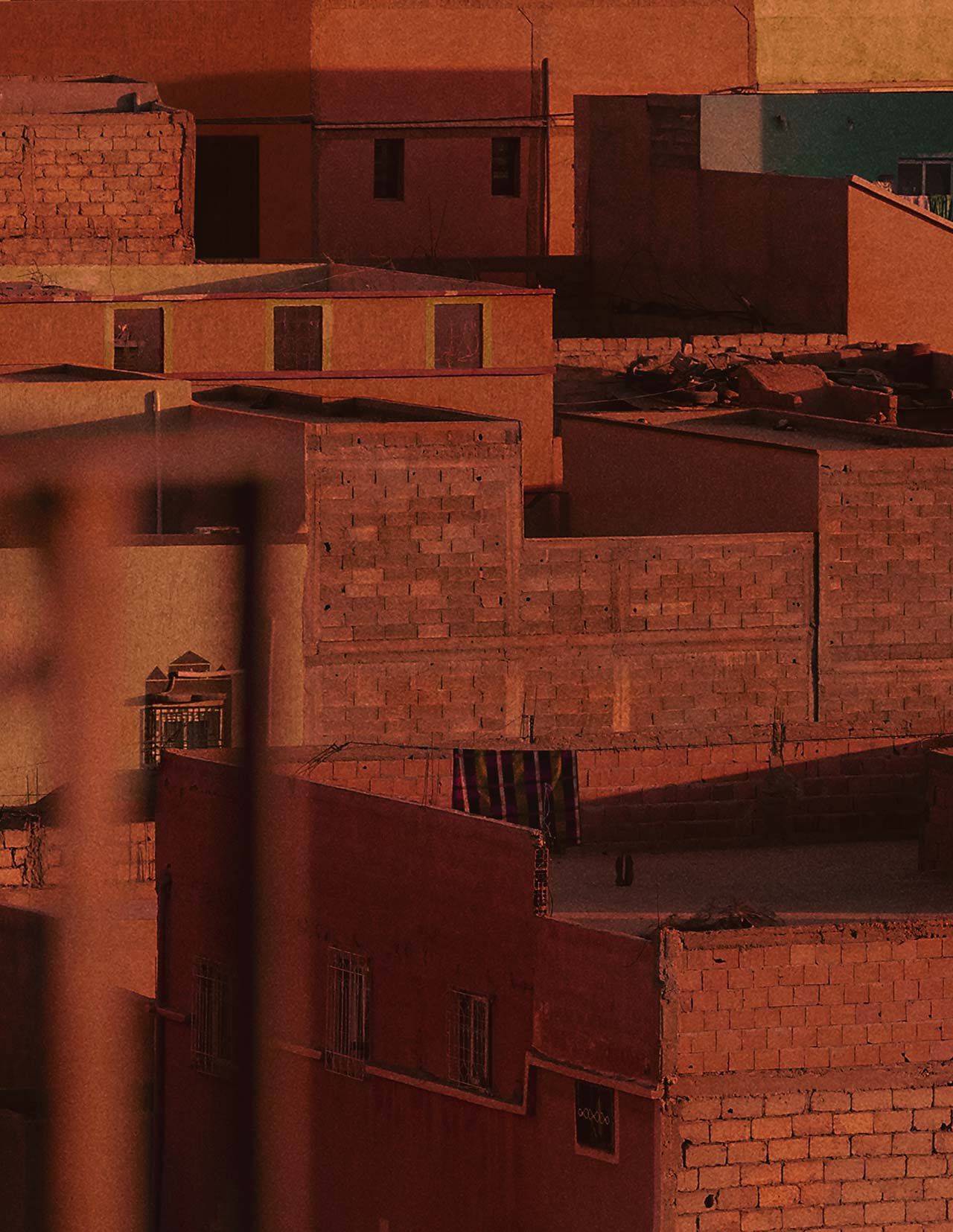 Tetti e mura cittadine color terracotta, avvolti da una calda luce rosso ruggine, con architetture squadrate, facciate in ombra e superfici murarie dalla texture marcata.