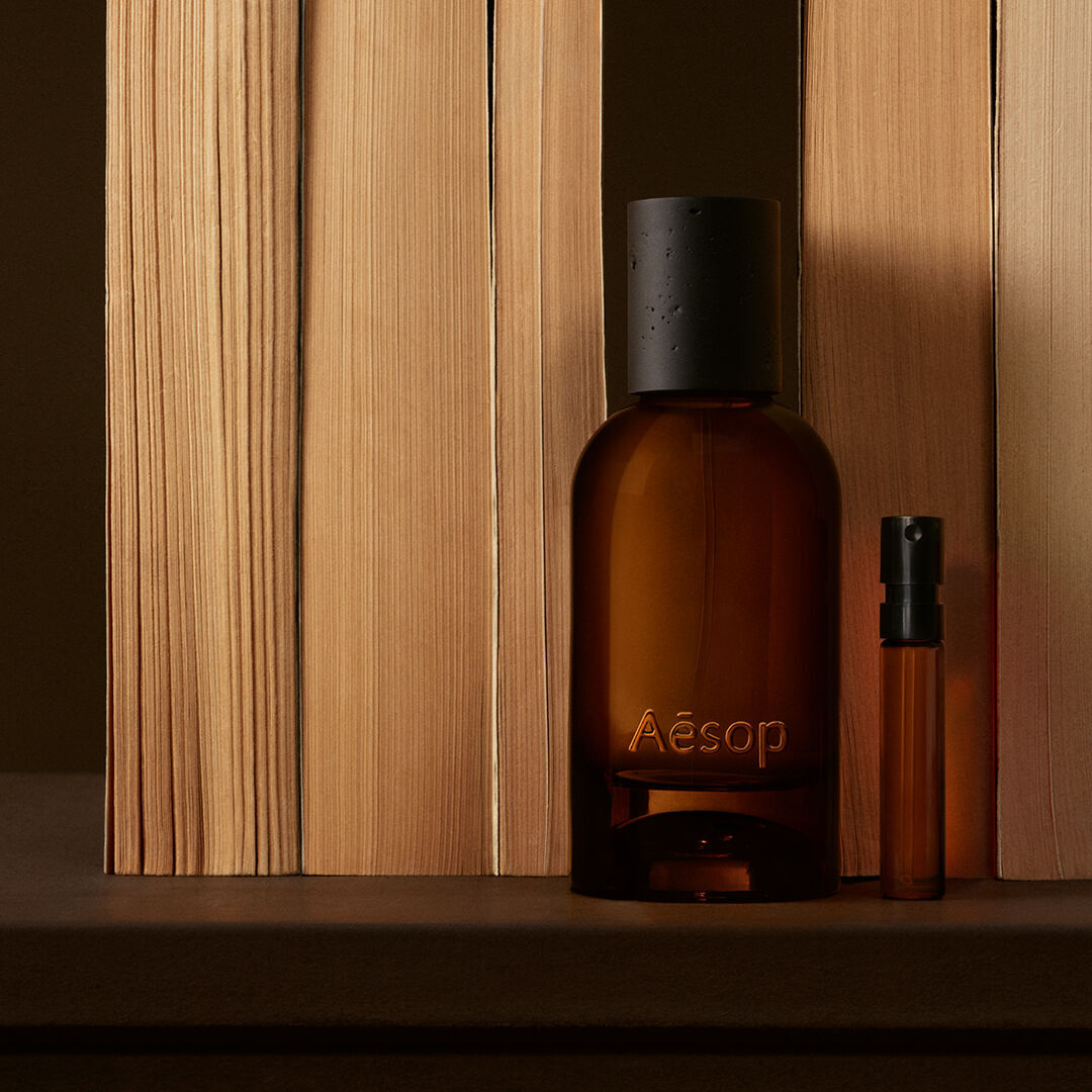 Flacone Aesop ambrato e fiala sottile disposti tra assi verticali di legno chiaro, con ombre bruno intenso e una composizione lineare, nitida.
