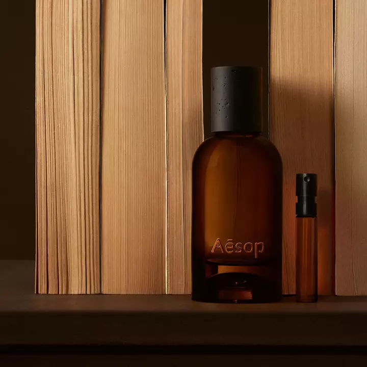 Flacone Aesop ambrato e fiala sottile disposti tra assi verticali di legno chiaro, con ombre bruno intenso e una composizione lineare, nitida.