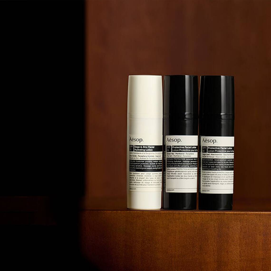 Un'accurata selezione di formulazioni per la protezione solare Aesop disposte su una superficie in legno scuro in un ambiente dalla luce soffusa.