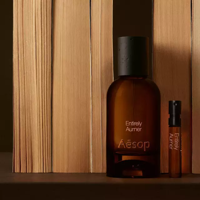 Un flacone di vetro ambrato del profumo Aesop Entirely Aurner e una fiala campione abbinata posizionati in piedi accanto a libri allineati verticalmente su una superficie scura.