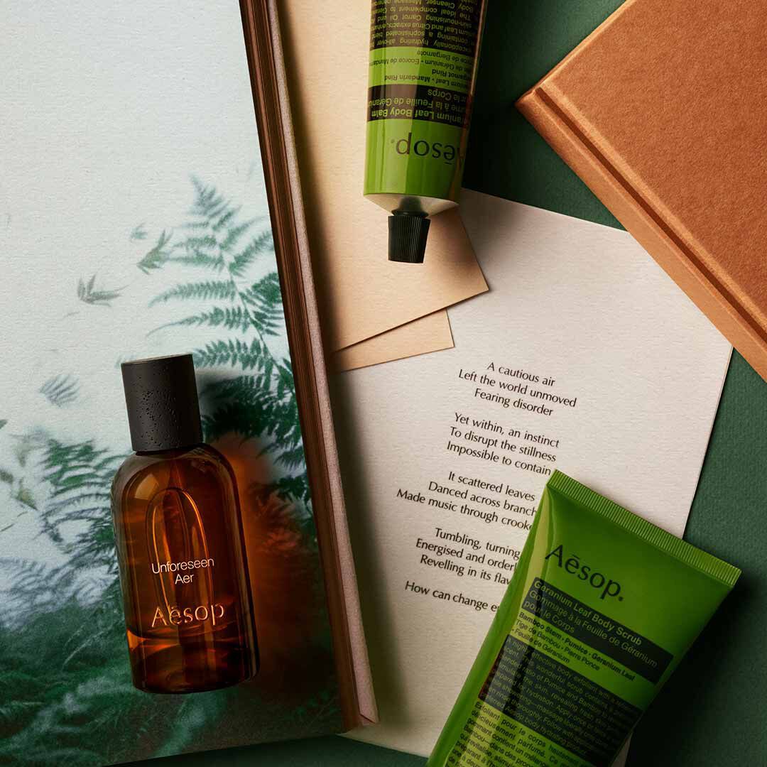 Flacone Aesop Unforeseen Aer con flacone a pompa e tubo verde, disposti su fogli con immagini di felci.