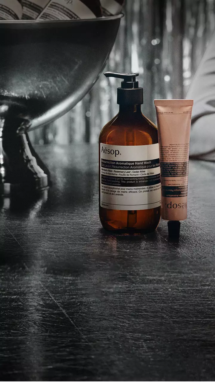 Un flacone ambrato Aesop con erogatore e un tubetto rosa si trovano accanto a una grande ciotola su piedistallo, su un tavolo riflettente, con festoni argentati sullo sfondo.