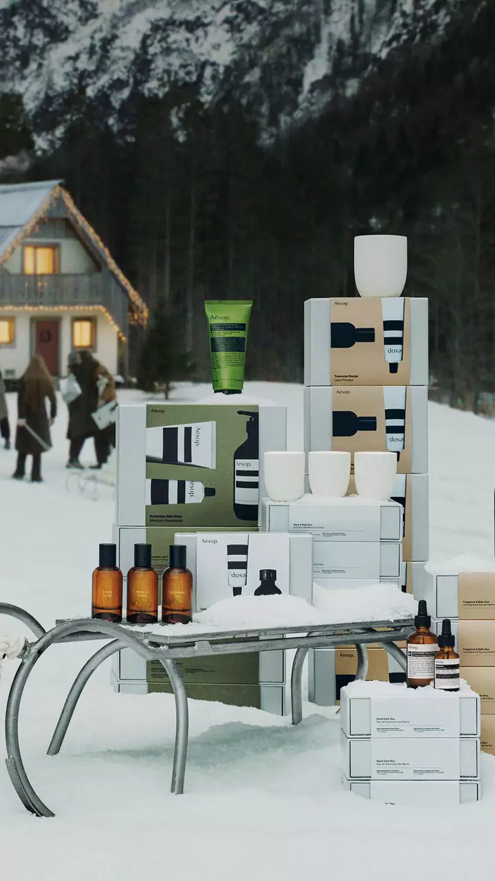 Scatole Aesop, flaconi ambrati, tubi colorati e recipienti bianchi sono disposti attorno a una slitta di metallo sulla neve, con persone all’esterno di una casa illuminata sullo sfondo.