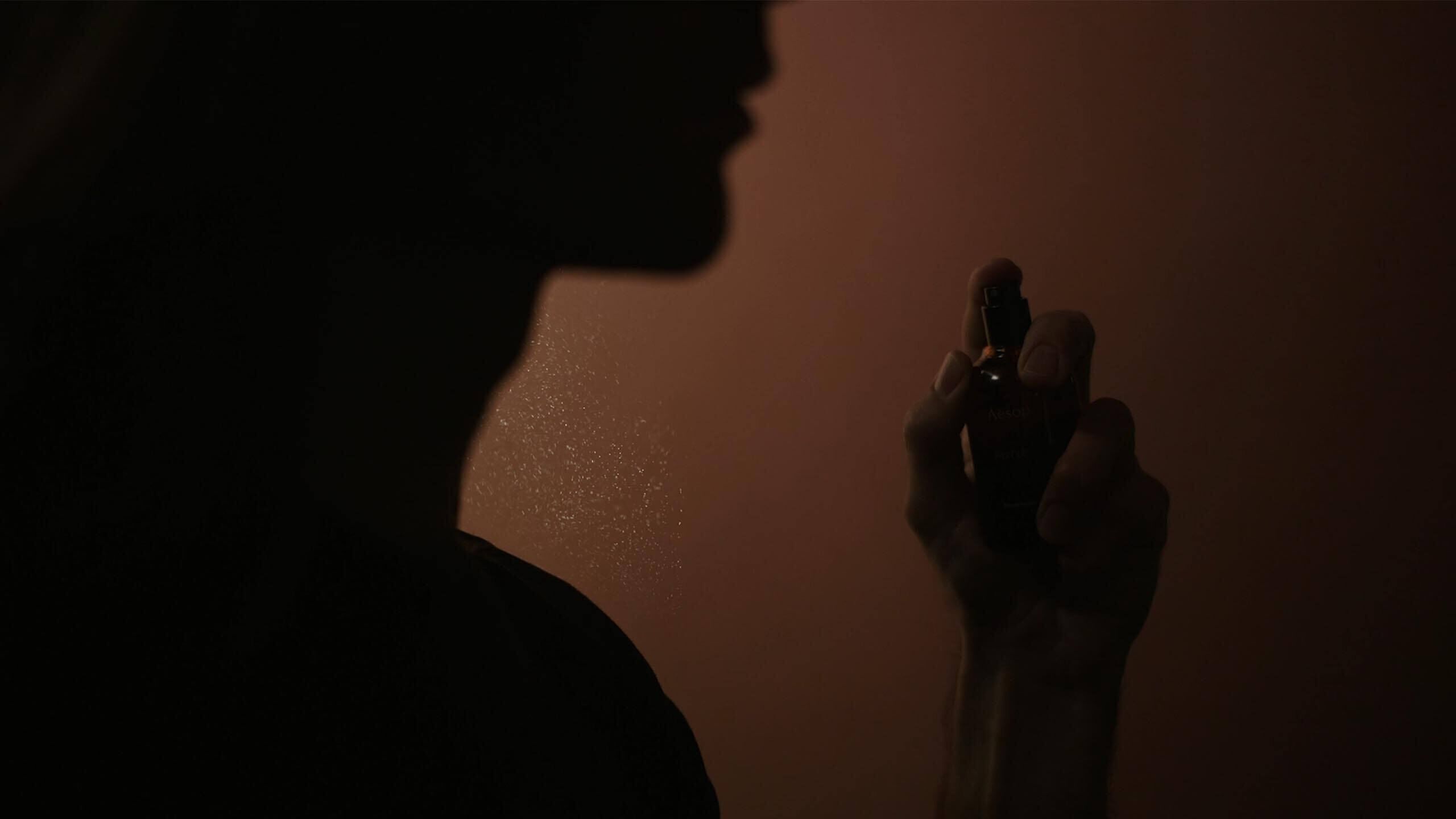 Silhouette di una persona che tiene una piccola boccetta di fragranza vicino al viso, su uno sfondo ambrato e caldo.