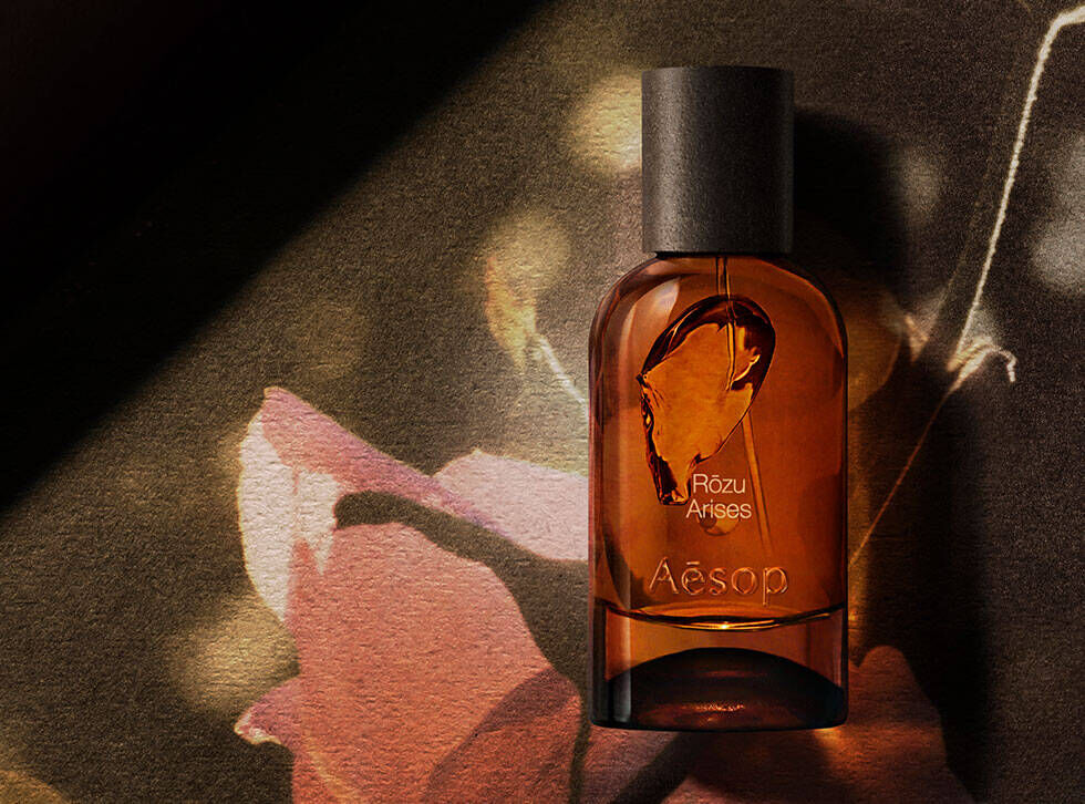 Flacone ambrato della fragranza Aesop Rozu Arises su una superficie scura, accanto a un’illustrazione botanica rosa e a una radice chiara.