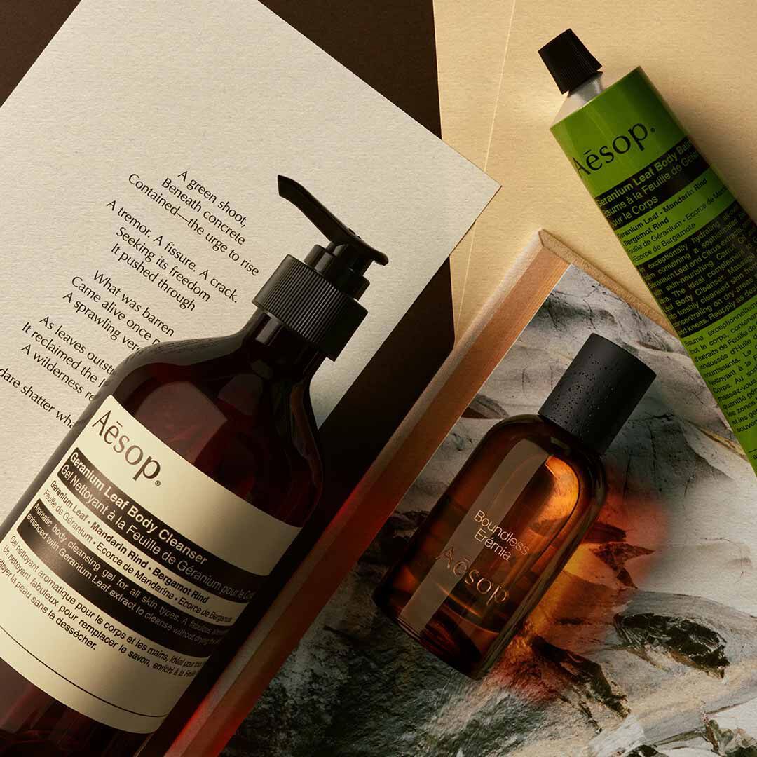 Flacone Aesop Boundless Eremia con flacone a pompa e tubo verde, disposti su fogli e un’immagine di pietra.
