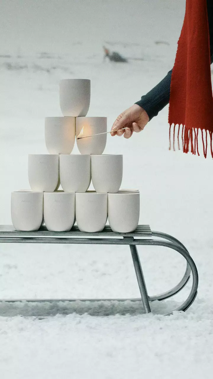 Una mano tiene in mano un fiammifero acceso accanto a recipienti bianchi impilati su una slitta di metallo nella neve, con una sciarpa rossa sfrangiata sulla destra.